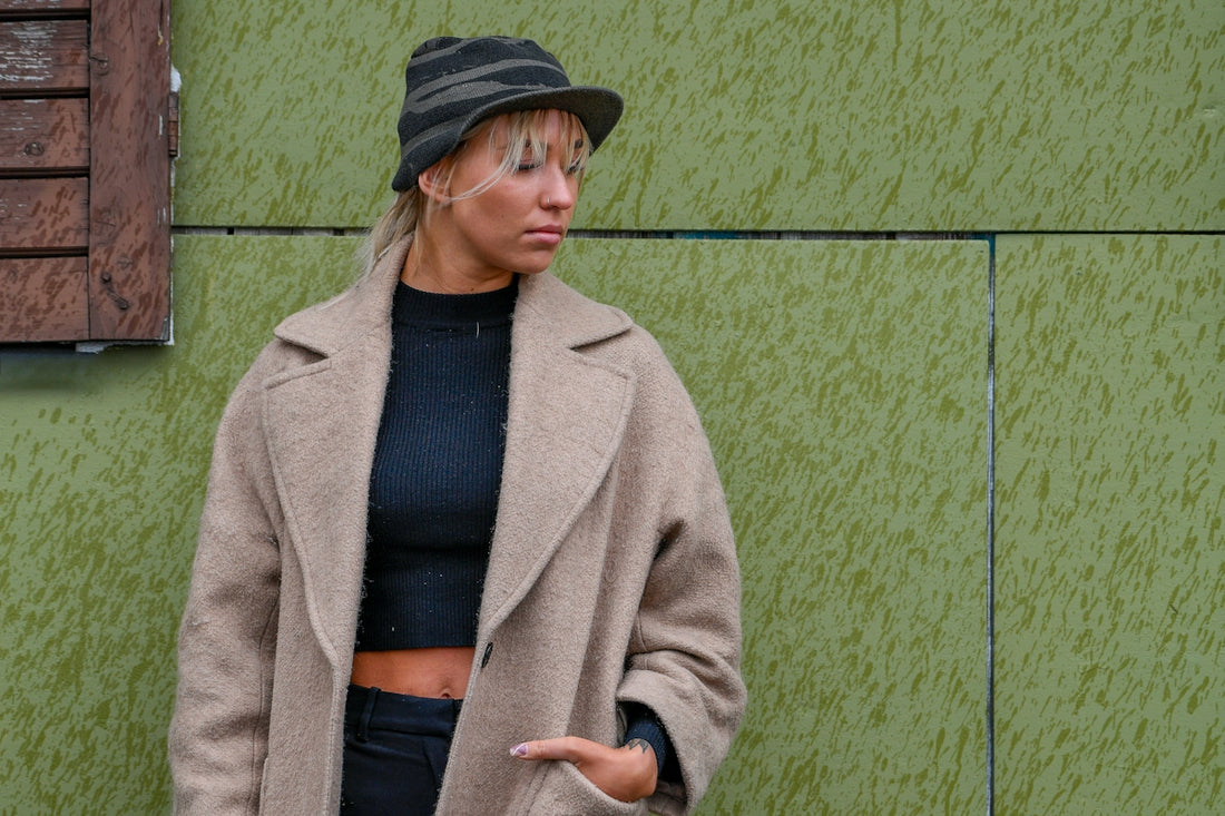 Woman in beige coat and striped hat against green wall.